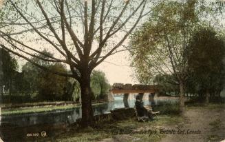 Colorized photograph of a man sitting on a park bench by the side of a river. In the background…