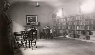 Picture of a room with tables and chairs and one wall of book shelves and tow women peaking int…