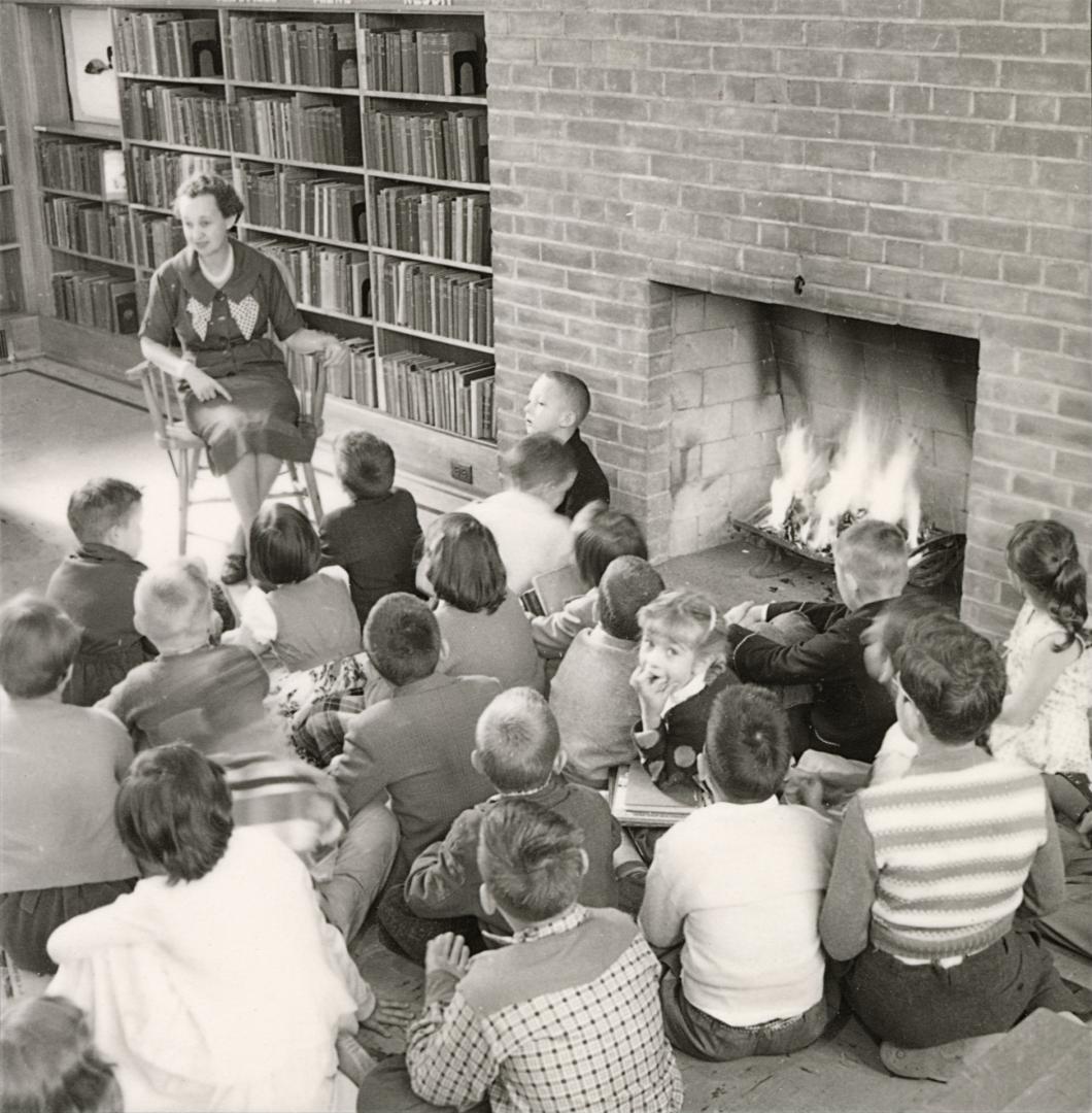 Picture of a group of children siting on the floor of a library in front of a blazing fire list…