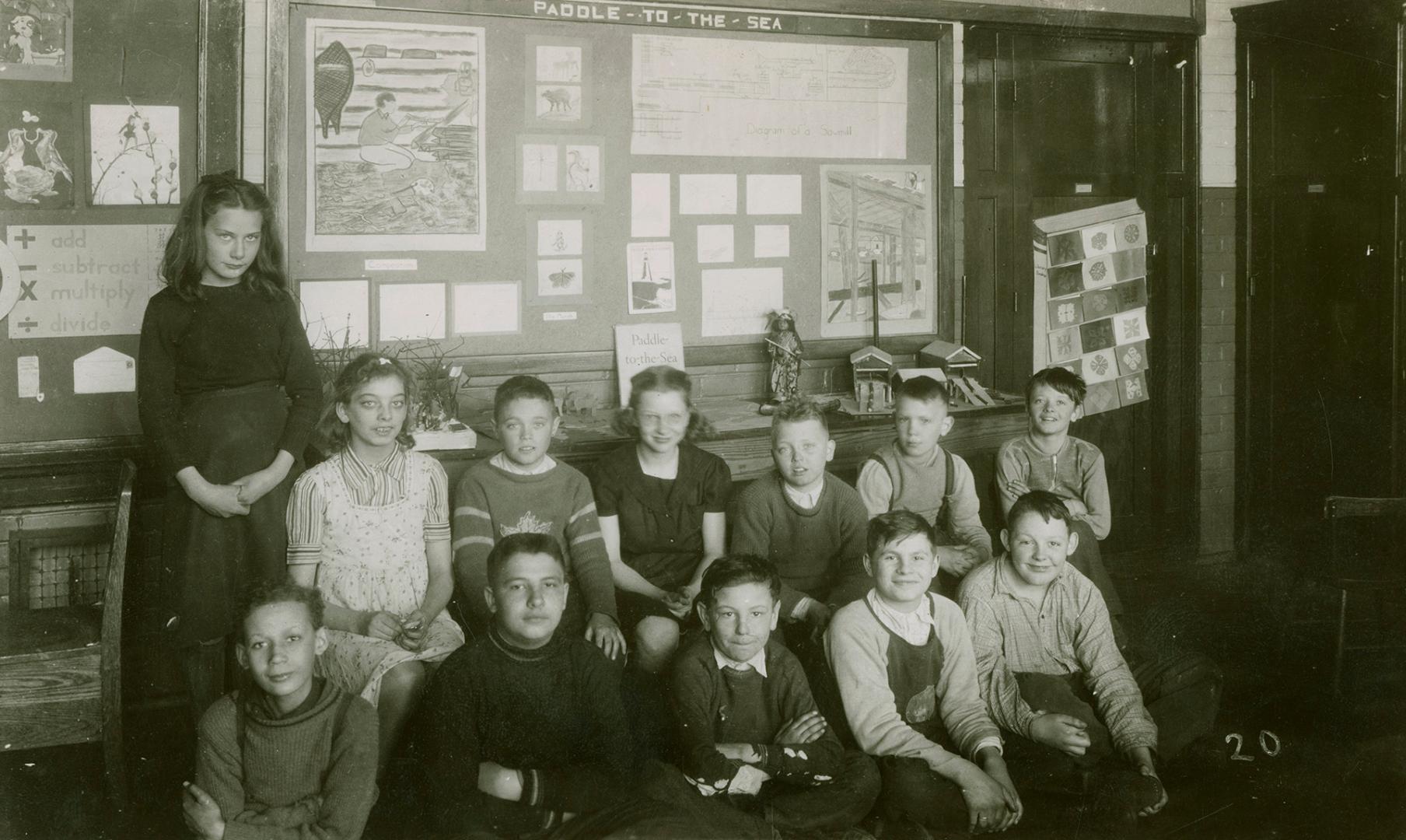 Picture of a group of children seated, with one standing, in front of a display board in the li…