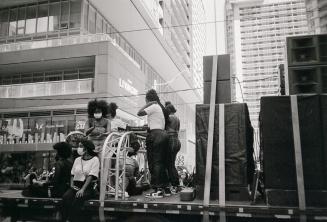 A group of people sit and stand on a flatbed truck parked in the middle of a street surrounded  ...