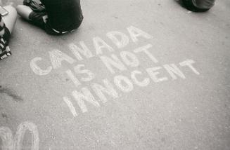 A photograph of a message reading &quot;CANADA IS NOT INNOCENT&quot; written on pavement in cha…