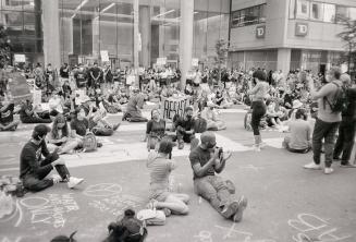 A photograph of a large group of people sitting and standing in the street and sidewalk in fron…