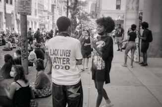 A photograph of several people sitting and standing on a road and sidewalk in front of a buildi ...