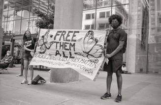 A photograph of two people standing in front of a police station holding a banner reading &quot…