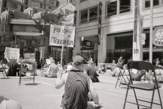 A photograph of several people sitting in the street and on the sidewalk in front of a police s ...