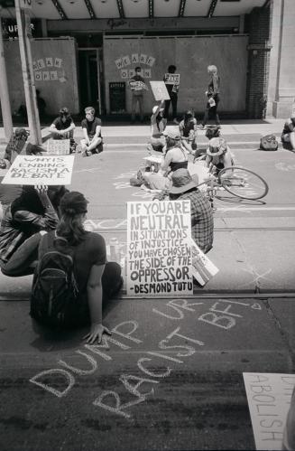 A number of people sitting on the street and on the sidewalk in front of a building. Some are h…