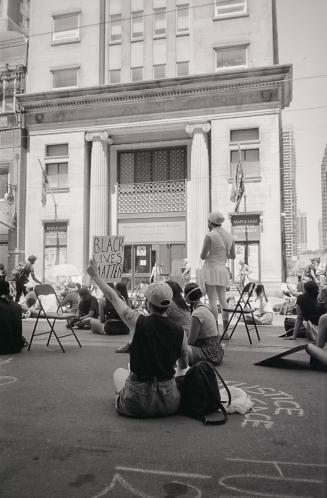 A photograph of a number of people sitting and standing on the road and sidewalk in front of a  ...