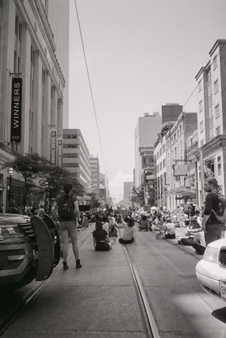 A photograph of a group of people sitting and standing in the middle of a street and on the sid…