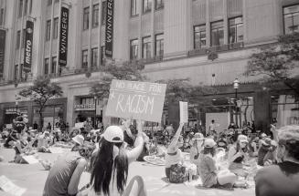 A photograph of a large group of people sitting in the middle of a road and on the sidewalk. Mo ...