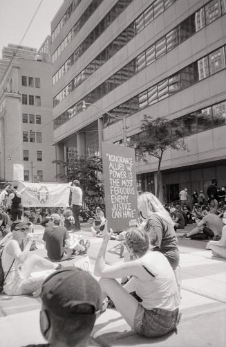 A photograph of a group of people, mostly young adults, sitting in the middle of a road and on  ...