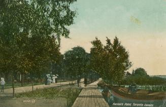 A boardwalk next to a sidewalk with people walking under large trees. 