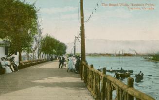 Picture of a wide boardwalk with people sitting and walking next to a lake with boaters. 