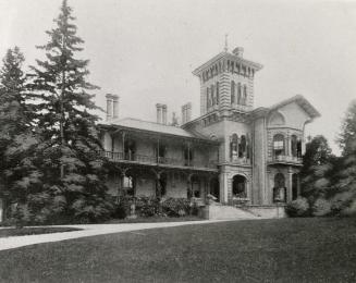 A photograph of a large two-story house, with third floor consisting of a tower. There are wide…