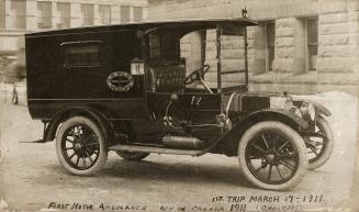 A photograph of a vintage ambulance parked in front of a building. There are no windows on eith ...