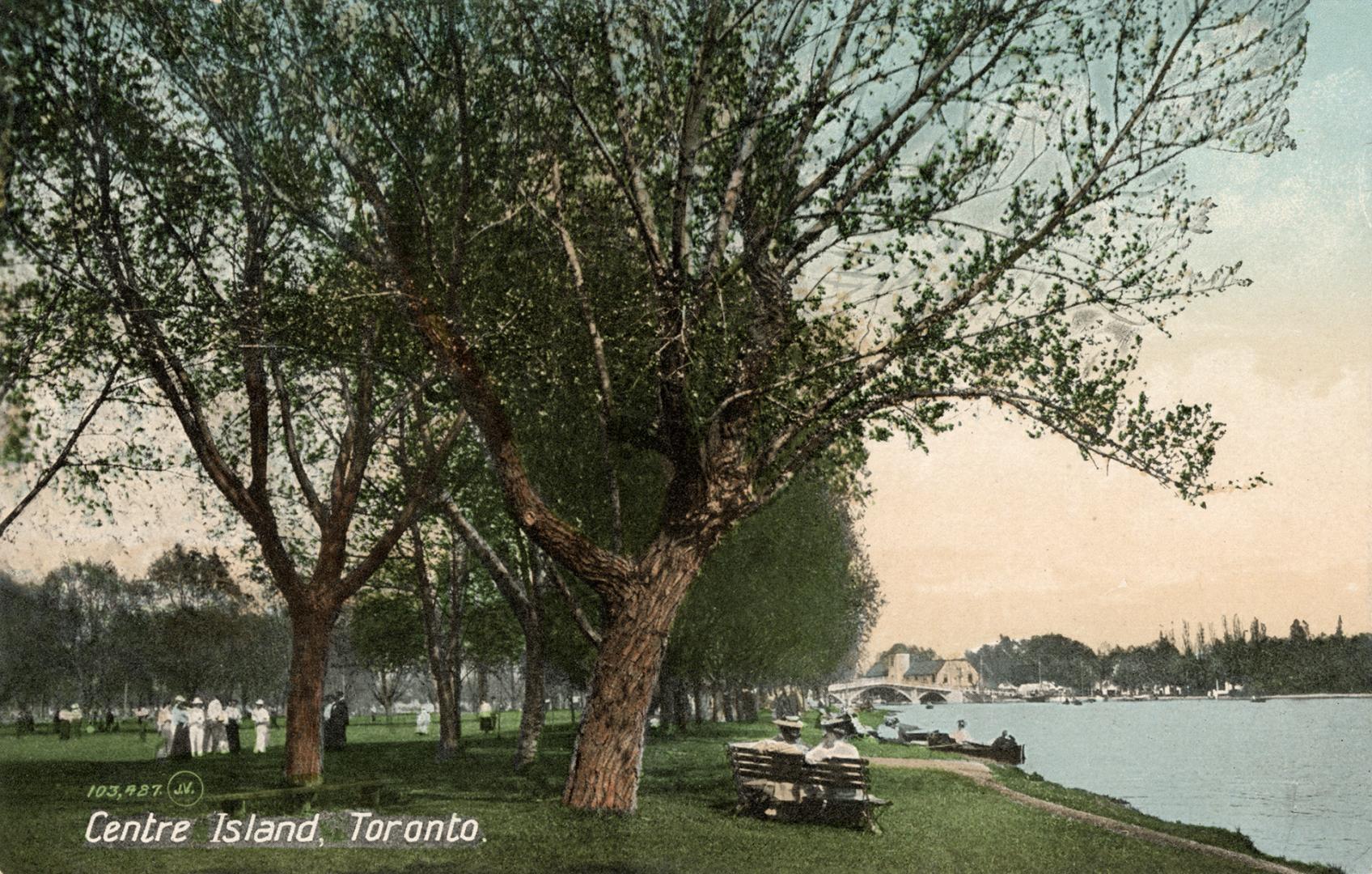  People enjoying the island on a sunny day under trees, beside lake with bridge in background.