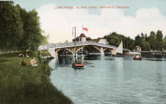 Picture of a bridge over a wide pond with boats in the water and people sitting on the grass at…