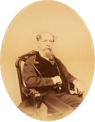 A photograph of an elderly man sitting in a wooden chair and facing the camera. He is wearing a…