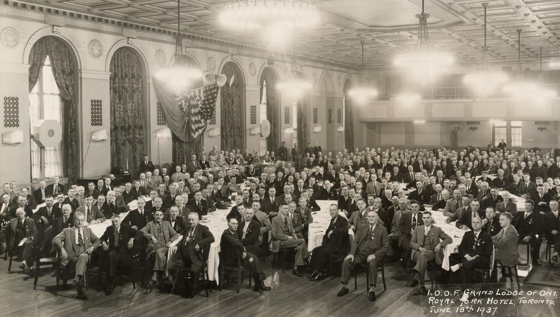 A photograph of a large group of men posing for a photograph while sitting in chairs around tab…