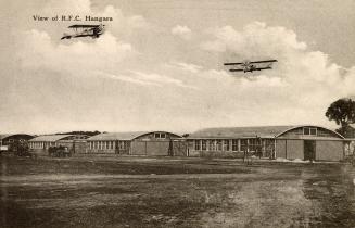 Sepia toned picture show two air planes flying over three low rise, commercial buildings.