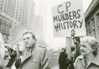 A photograph of a group of people holding picket signs in front of a building. On the left a mi…