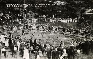 Black and white photograph of crowds of people standing around and swimming in a huge, public p…