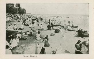 A photograph of a public beach area, with a large number of people swimming and sitting on the …