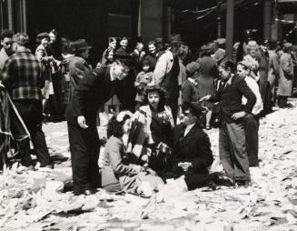 A photograph of a group of people sitting and standing in the middle of a city street which is …