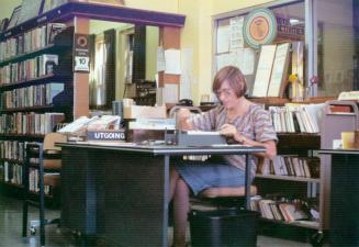 Picture of a librarian sitting at a check out desk surrounded by book shelves. 