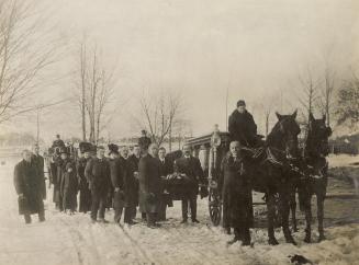 A photograph of a funeral procession. At the front of the procession there is a wagon being pul…