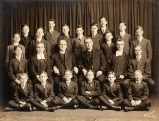 A formal class photograph, taken in a studio, of twenty-four students and teachers. The student…