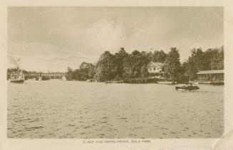 Black and white photograph of a motor boat on a lake with cottages on the shoreline. Bridge in …