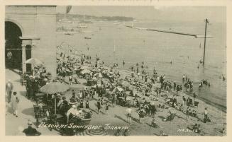 Crowds of people on a beach at a lake with a building in the left foreground. 