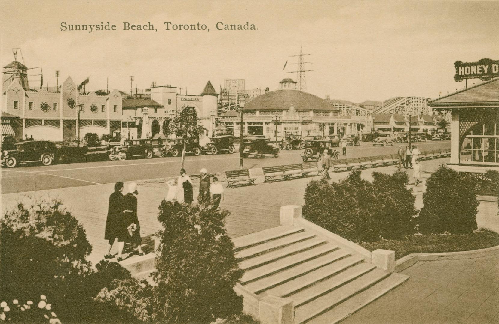 People walk on a boardwalk with stairs and bushes on the right and a road and amusement park in…