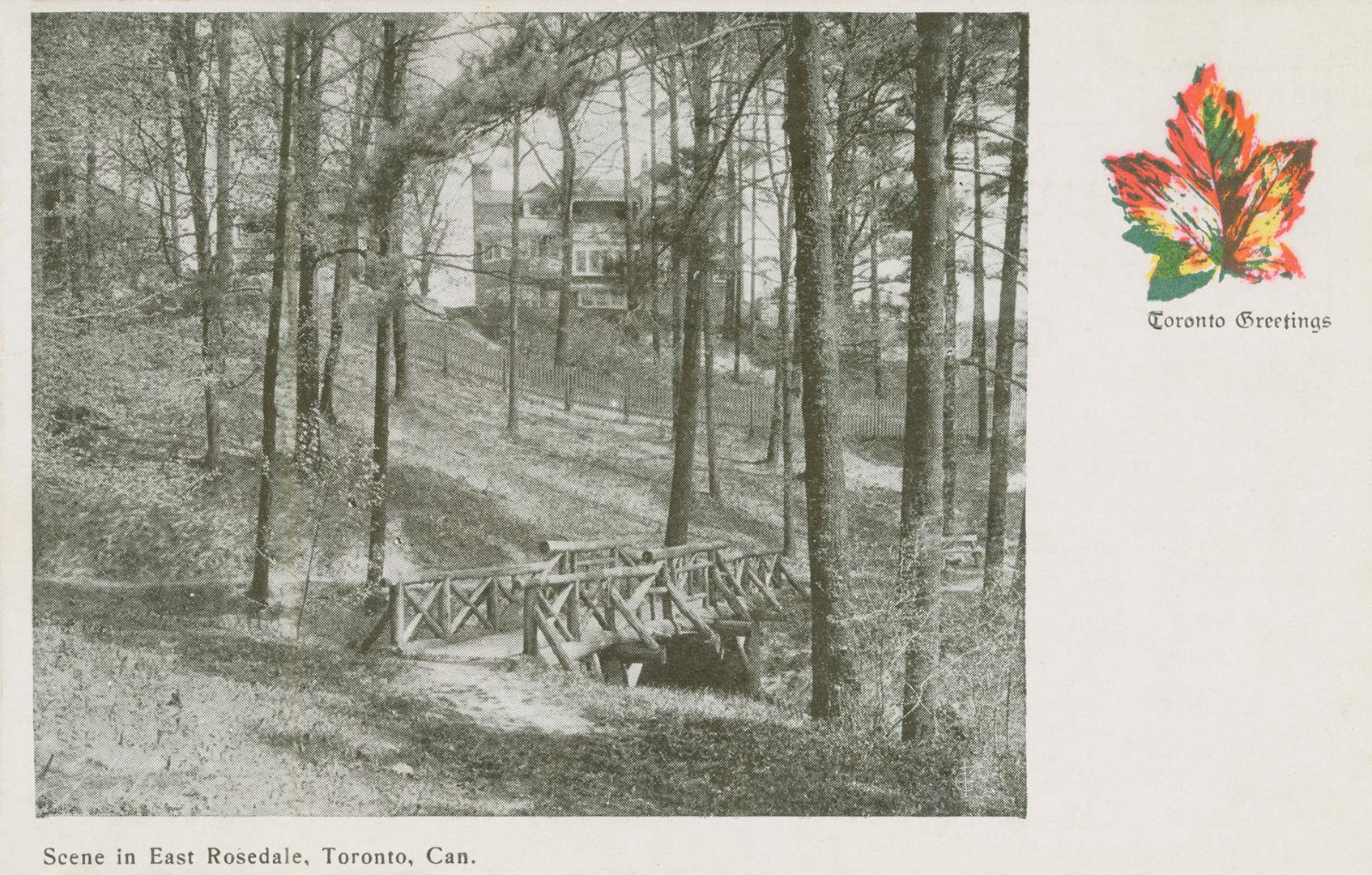 Black and white picture of a log bridge in a wooded area with large houses in the background.