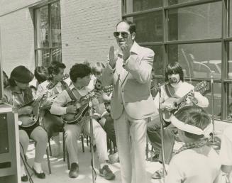 A group of young musicians seated outdoors are led by the music director who is clapping along.…