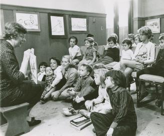 A librarian reads a story book to a group of children seated in front of her on the floor and o…