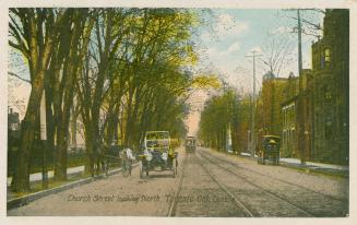 Street car and cars sun along a city street lined with trees on the left and buildings on the r…