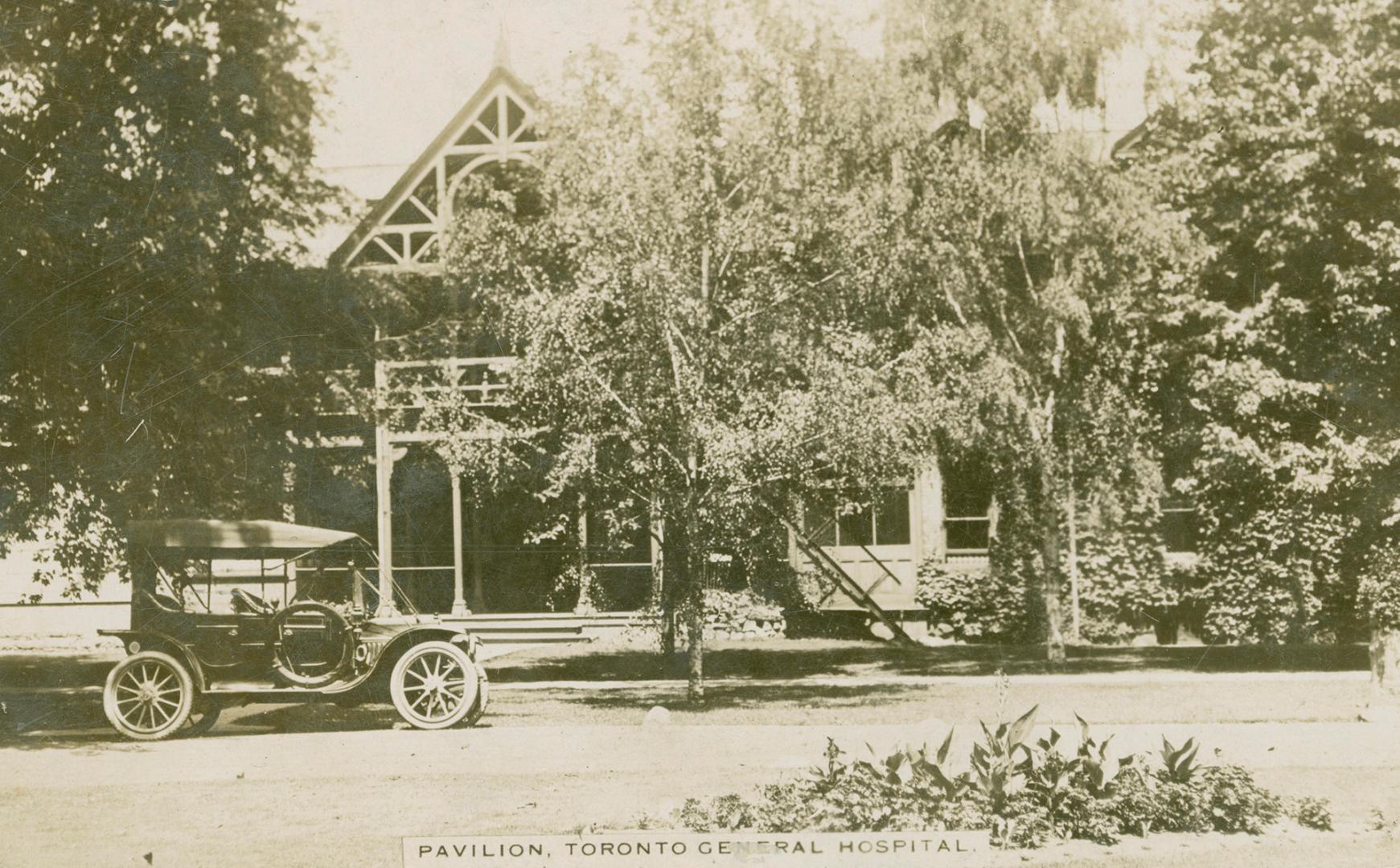 Black and white picture of a car parked in front of a two story wooden building with trees in f…