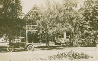 Black and white picture of a car parked in front of a two story wooden building with trees in f…