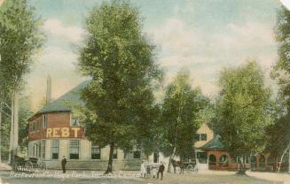 People and horse drawn carriages in front of a two story commercial building in a wooded area.