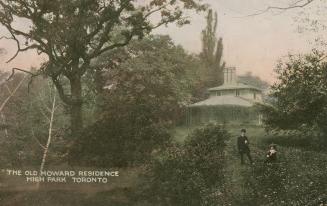 Two people standing and sitting in front of two story home in the middle of a wooded area.