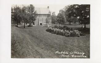 Picture of a park with flower beds and trees and public library in background. 