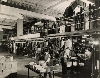 A photograph of a a large printing press, with several people working at and around it. In the …