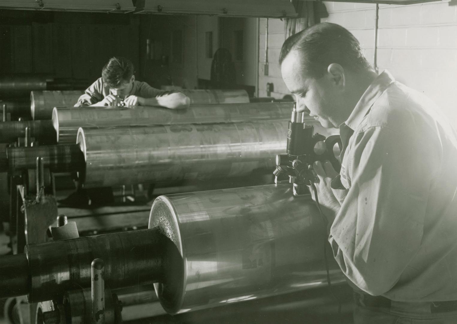 A photograph of two men using microscopic devices to examine images on cylinders during the pro…