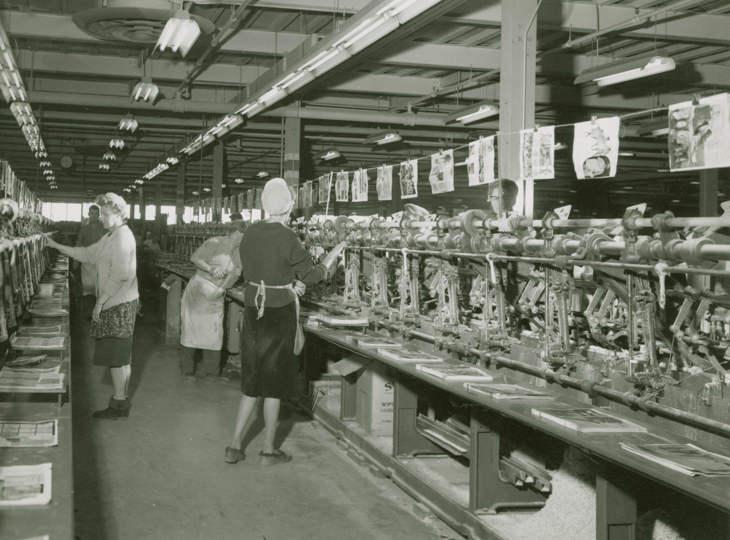 A photograph of men and women in front of a metal assembly line, with piles of pages of catalog…