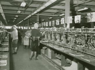 A photograph of men and women in front of a metal assembly line, with piles of pages of catalog…