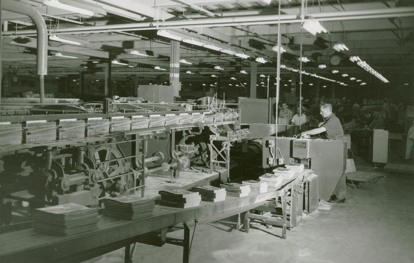 A photograph of a man standing at the end of a moving assembly line, with piles of Simpsons-Sea…