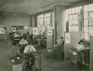 A photograph of an office with warehouse-style windows on the right side and men and women work…