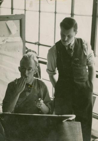 A photograph of two men, one sitting and one standing, in front of a drafting table. The standi…